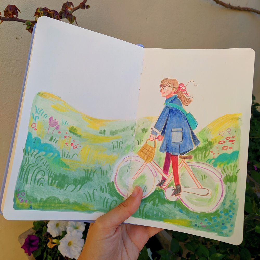 photo of my hand holding my sketchbook. I painted a girl riding a bike through a peaceful green grass field.