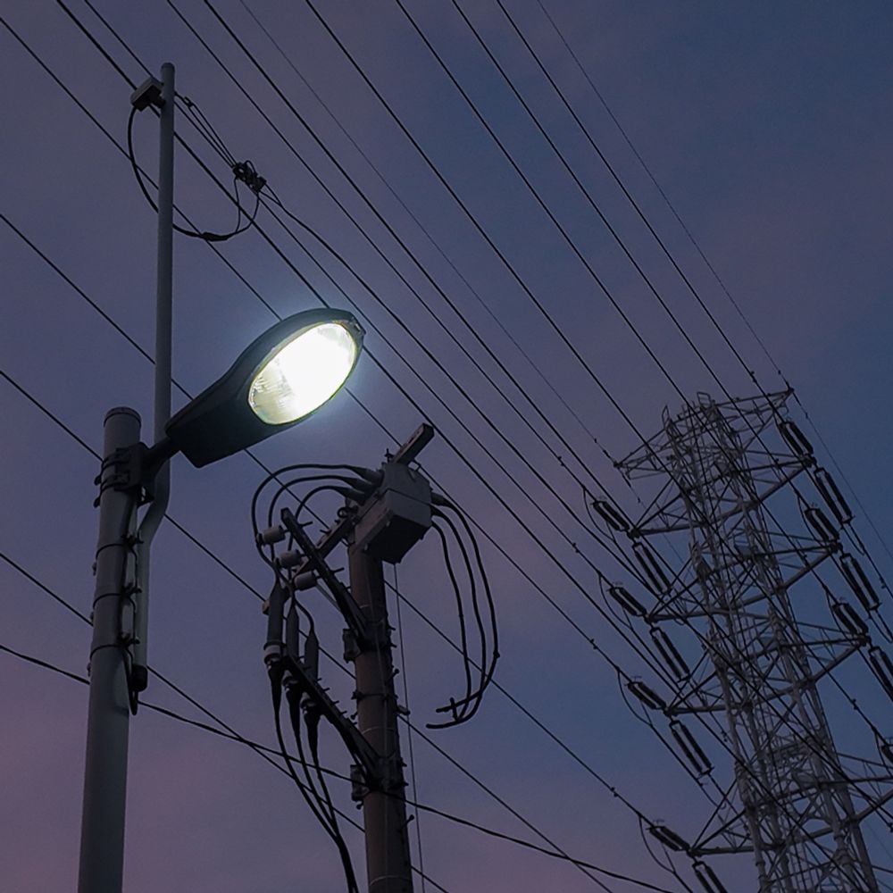 夕暮れの空を背景に、街灯と鉄塔が静かに光を灯して立っている様子。 A streetlight and a transmission tower quietly standing and glowing against a dusk sky.