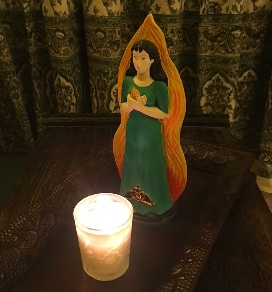 Photograph of a votive candle set before a statue of a person with long black hair, white skin, and a green dress, holding a small flame.  The lit candle illuminates the statue which is backed by red-yellow fire.  Both sit on a bronze tray before a curtain with green and purple designs.