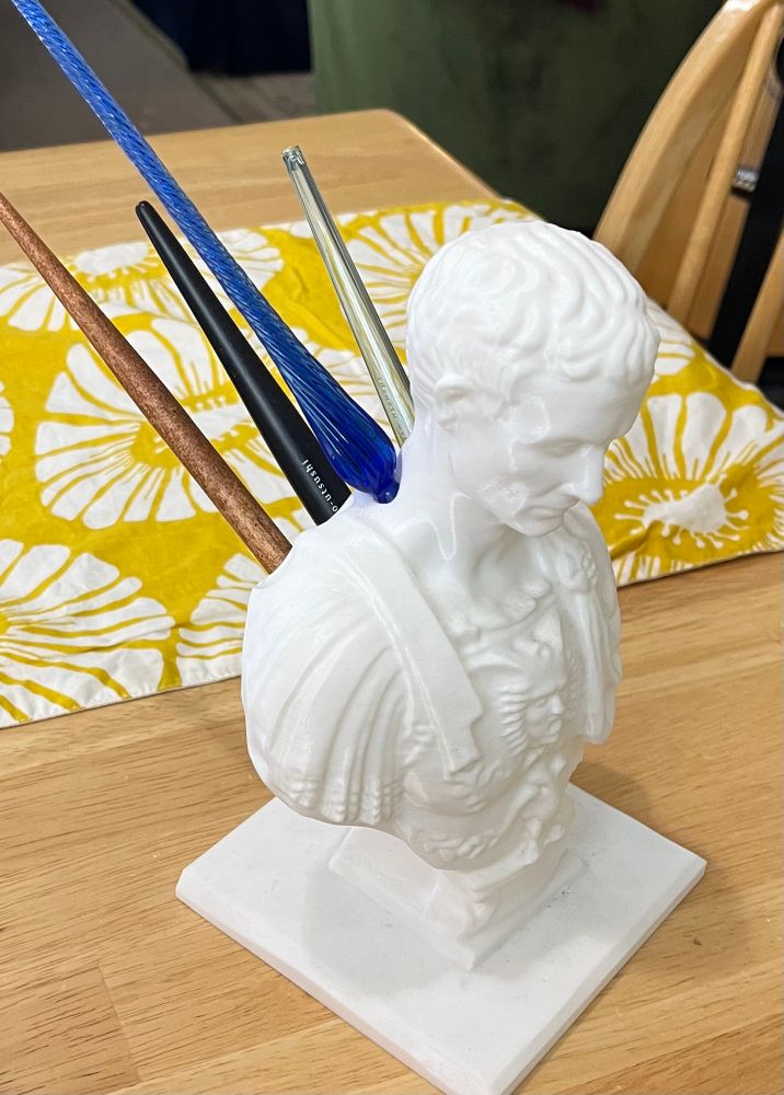 A white plastic bust of Caesar about 8 inches high sitting on a wooden table with a yellow and white floral table runner. The bust is actually a pen holder, and it has four calligraphy pens poking from its back in the colors of black, clear, dark blue, and wood tone. 