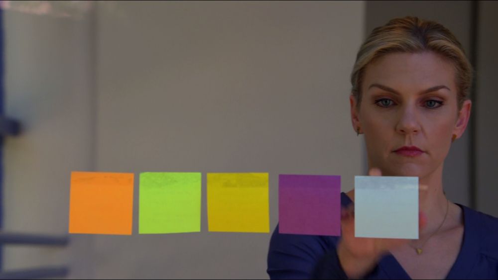 Kim Wexler hangs an orange, a green, a yellow, a purple, and a blue post-it notes on a window.