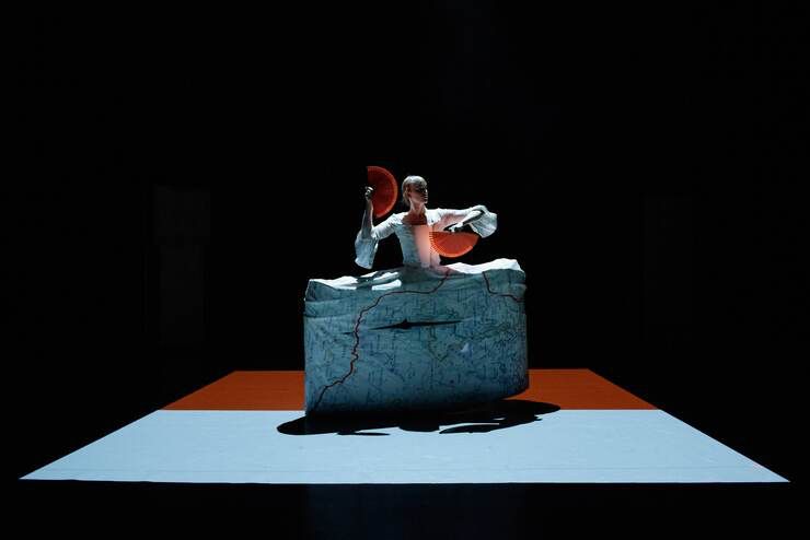 A performer stands in a black box room on a white and orange square flooring. They’re wearing a custom dress in the design of a map and holding fans in both hands.