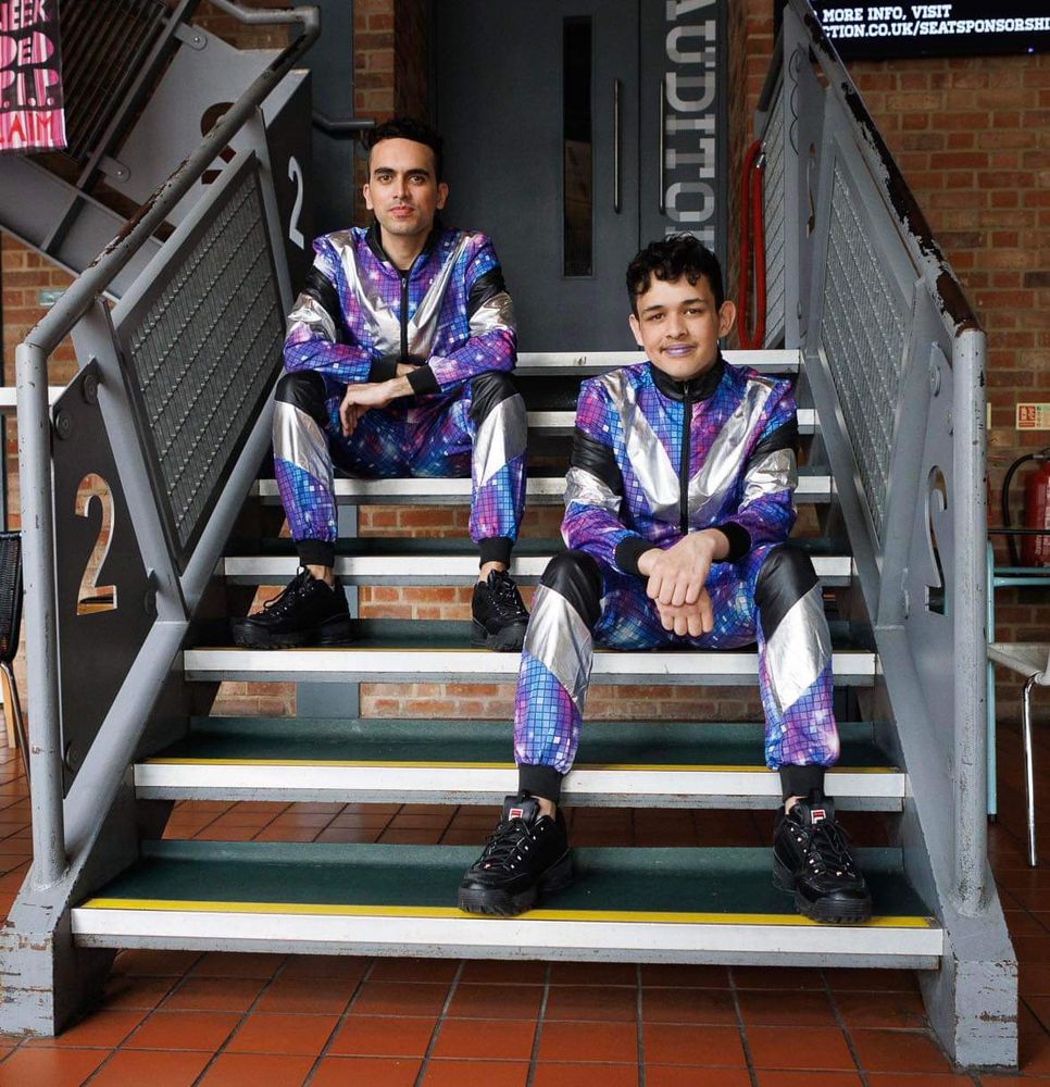 Two brown, autistic brothers sat on a metal staircase wearing two piece matching disco tracksuits and black trainers. The image looks very much like an album cover for a boy band.