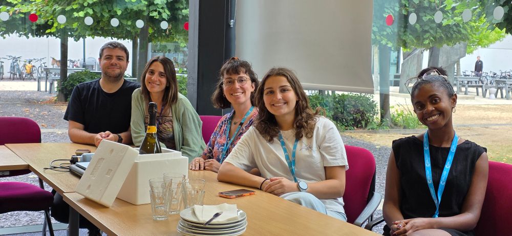Pathania lab team members sitting at a desk waiting for prosecco and some cake to be served up 