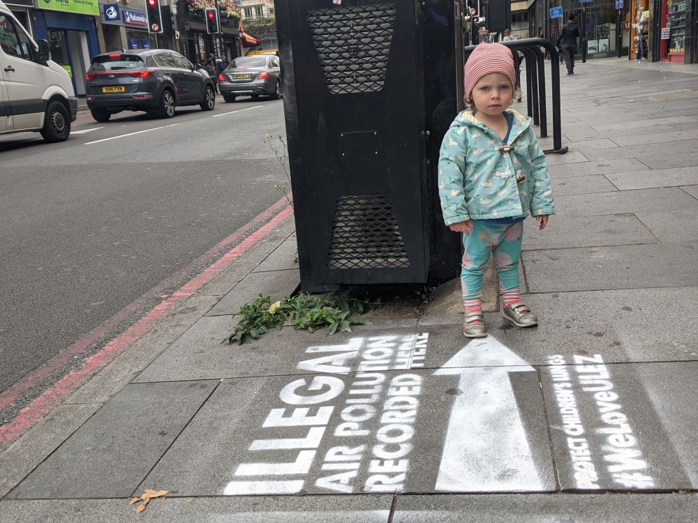 A child stands in front of the Brixton Road air pollution monitor. A chalked stencil on the ground says "Illegal air pollution recorded here". 