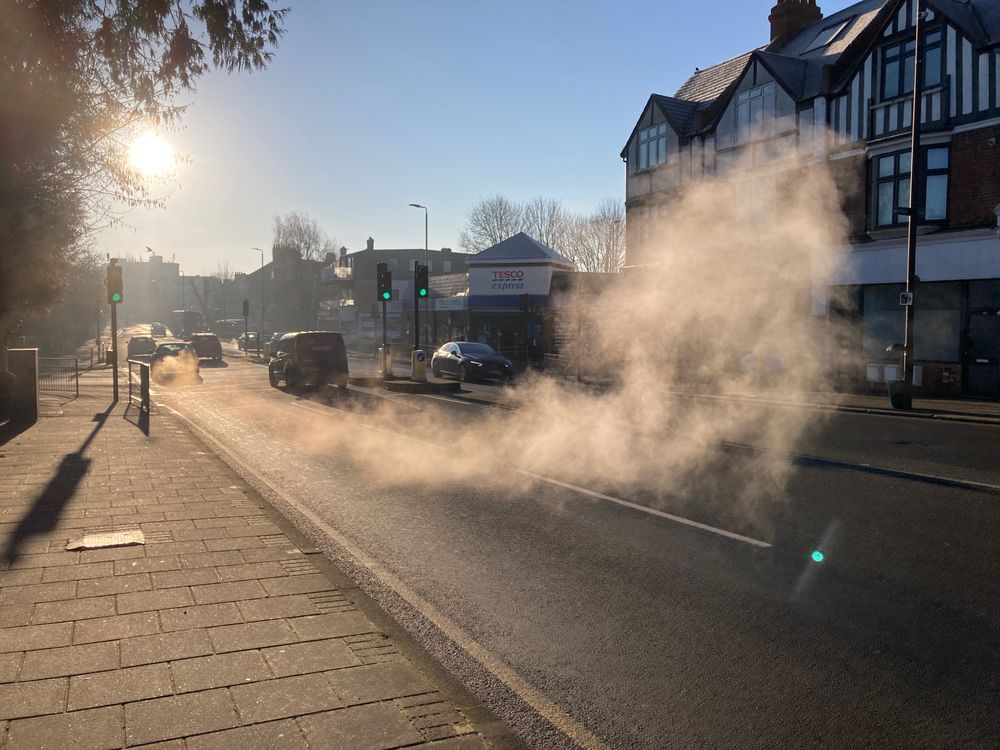 A road with some cars travelling. One has a very smoky exhaust and the fumes have stayed in the air after is has accelerated away. 
