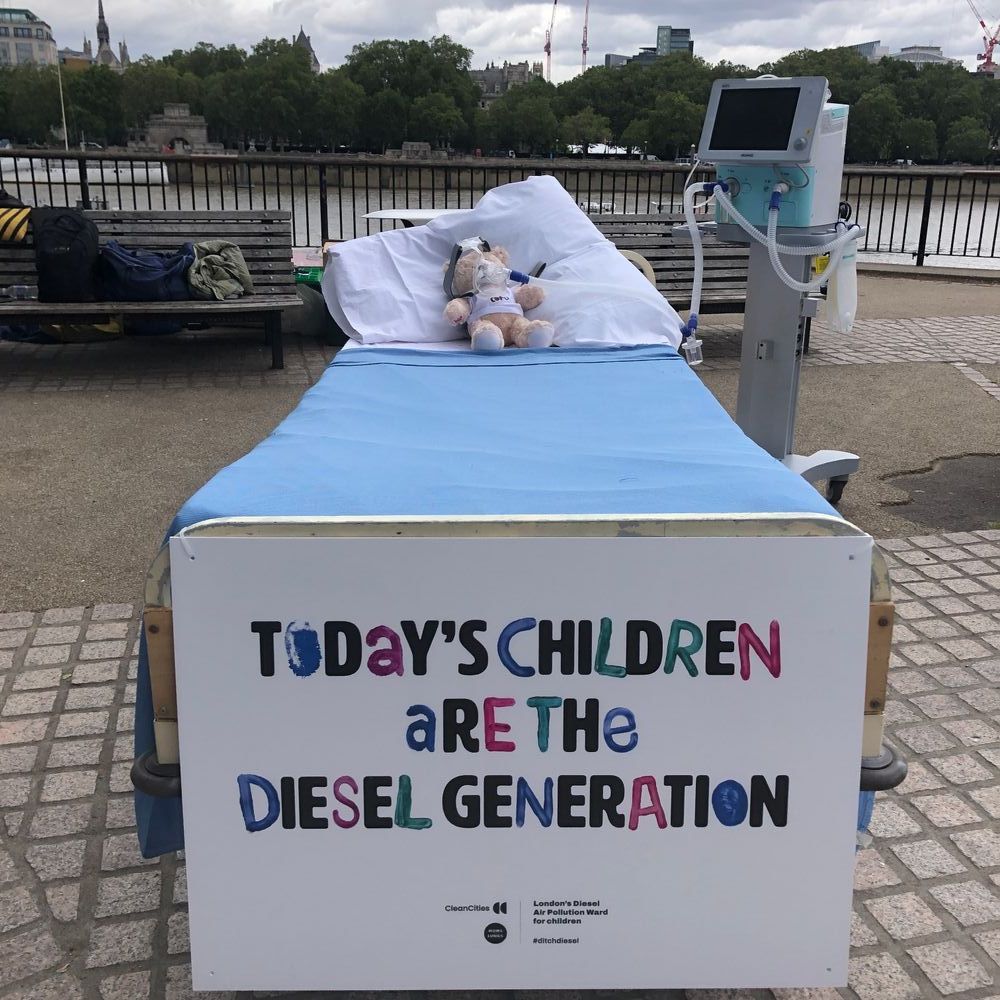 A hospital bed on London's South Bank with a teddy in it, hooked up to a machine. The placard on the bed says "Today's children are the diesel generation". 