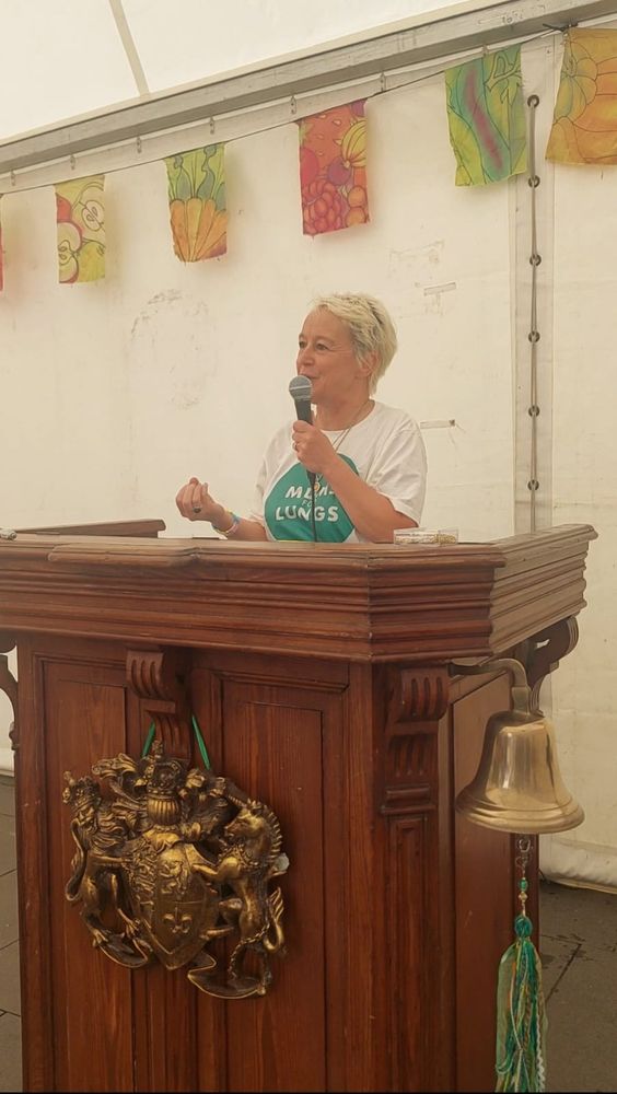A woman with a microphone, giving a talk, standing behind a large wooden podium with a brass crest on the front. 