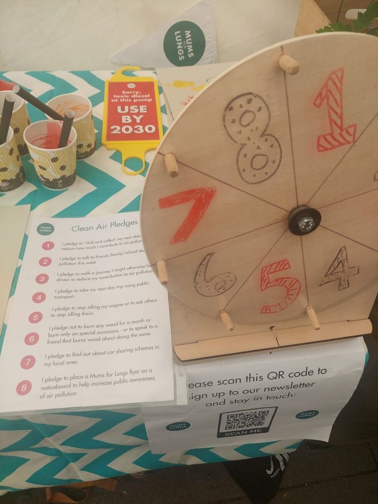 Part of a table with a green and white tablecloth on it. On the table are some air pollution campaigning resources - some clean air pledges and a wooden spinning wheel to select a pledge. 