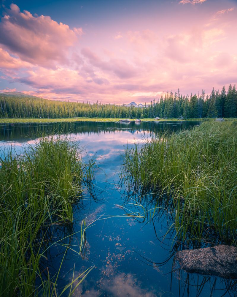Looking out over a mountain lake towards a distant peak as the sun sets. The clouds and trees are reflected in the still water.