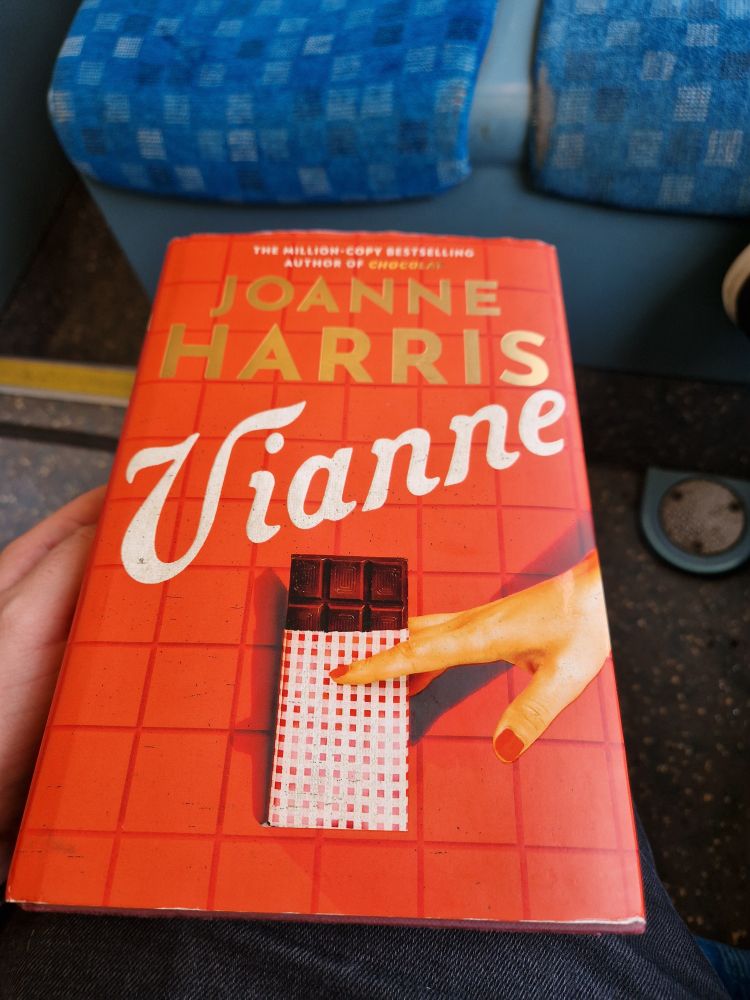A copy of "Vianne" by Joanne Harris resting on a knee. The cover is orange with dark crosses like a tablecloth. In the centre is a bar of dark chocolate, pincered between fingers painted with red nail barnish.