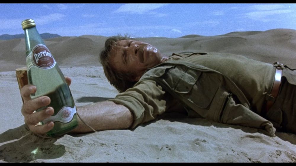 Chuck attaché dans le désert, une bouteille de Perrier à la main, l'étiquette bien tourné vers la caméra 