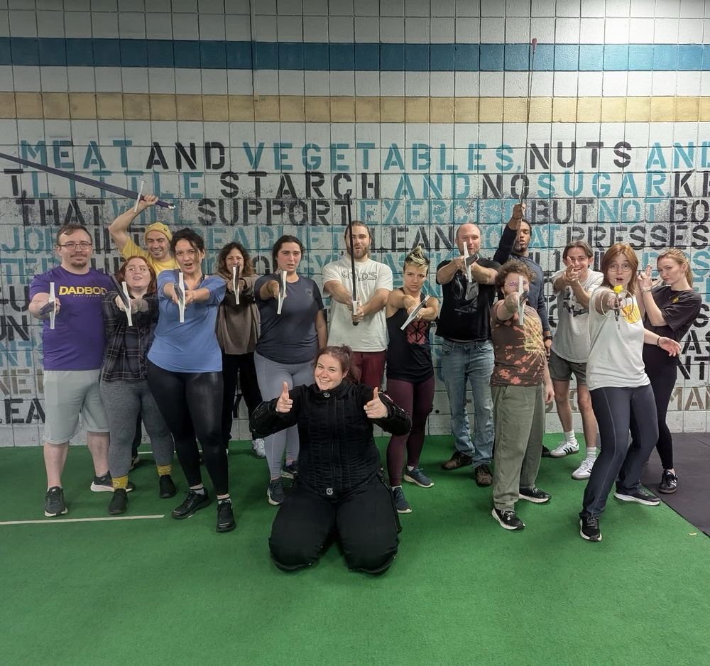 A group of people in a gym. They are holding swords towards the camera. 
