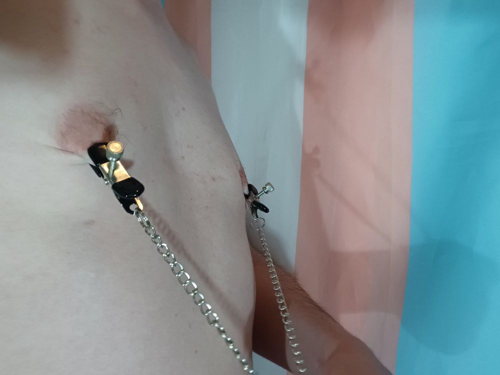 A man stood shirtless in front of a trans flag. He is wearing nipple clamps attached by a chain, which is being pulled downwards. 