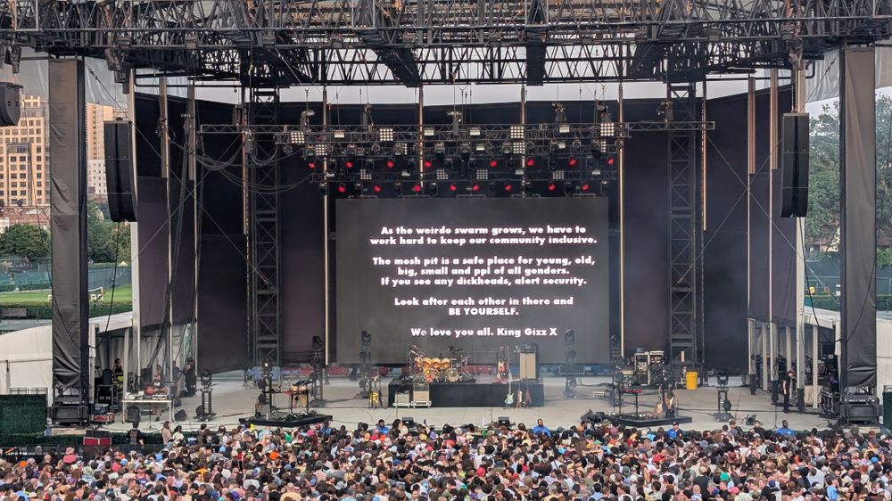 A large crowd is gathered in front of an outdoor concert stage. A large screen at the back of the stage displays the following text:

As the weirdo swarm grows, we have to work hard to keep our community inclusive.

The mosh pit is a safe place for young, old, big, small and ppl of all genders. If you see any dickheads, alert security.

Look after each other in there and BE YOURSELF.

We love you all. King Gizz X