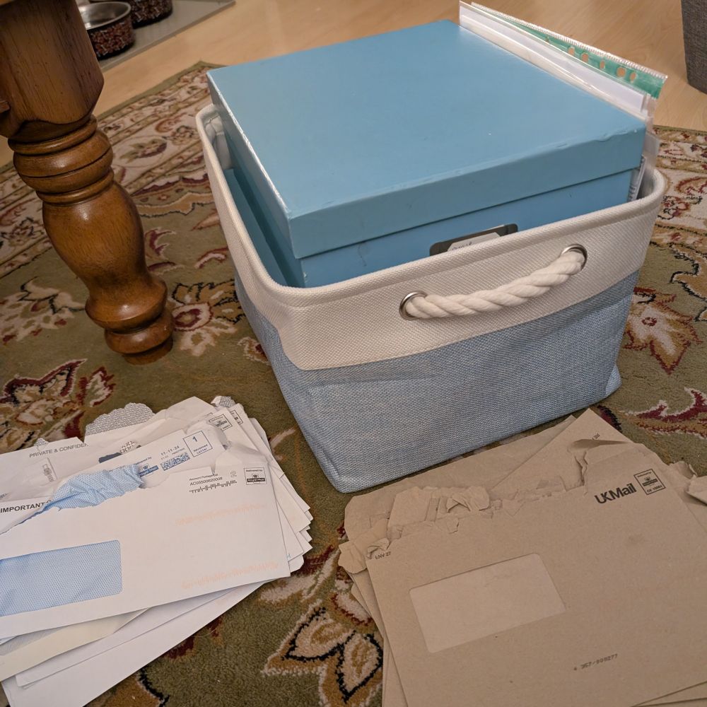 An photogaph showing a pile of empty white and brown envelopes on the floor in front of a white and blue canvas storage box with organisational file boxes and plastic pockets overflowing over the top of the storage box.