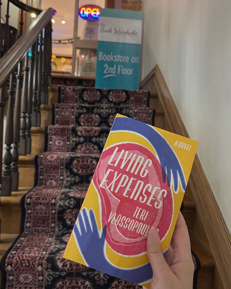 My hand holding a copy of Teri Vlassopoulos’ novel Living Expenses, with a staircase in the background and a sign that reads “The Book Wardrobe, Bookstore on the 2nd floor,” with an Open sign on the shop window.