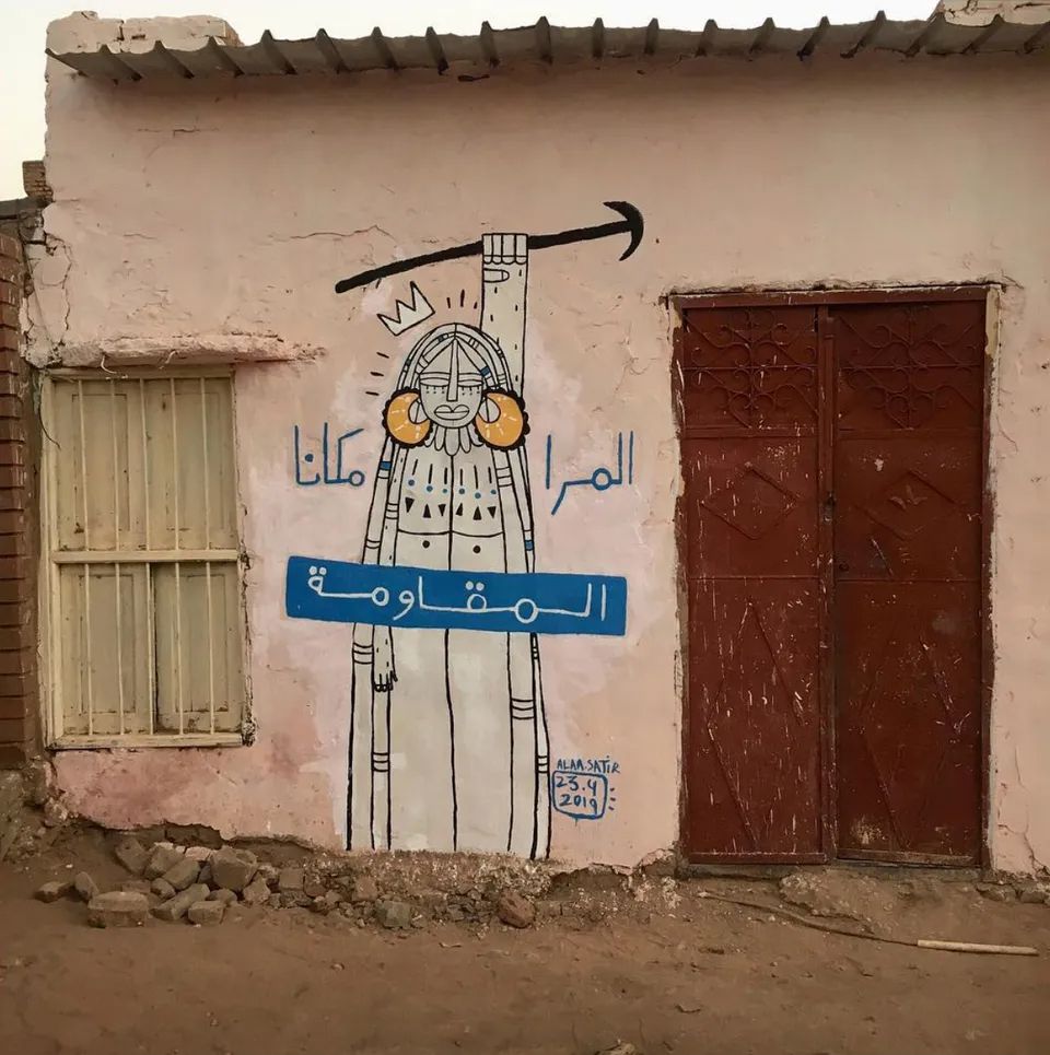 Artwork on a residential wall in Khartoum of a woman holding up a stick usually used for cooking. The text says A woman's place is the resistance in Arabic
المرا مكانا المقاومة.