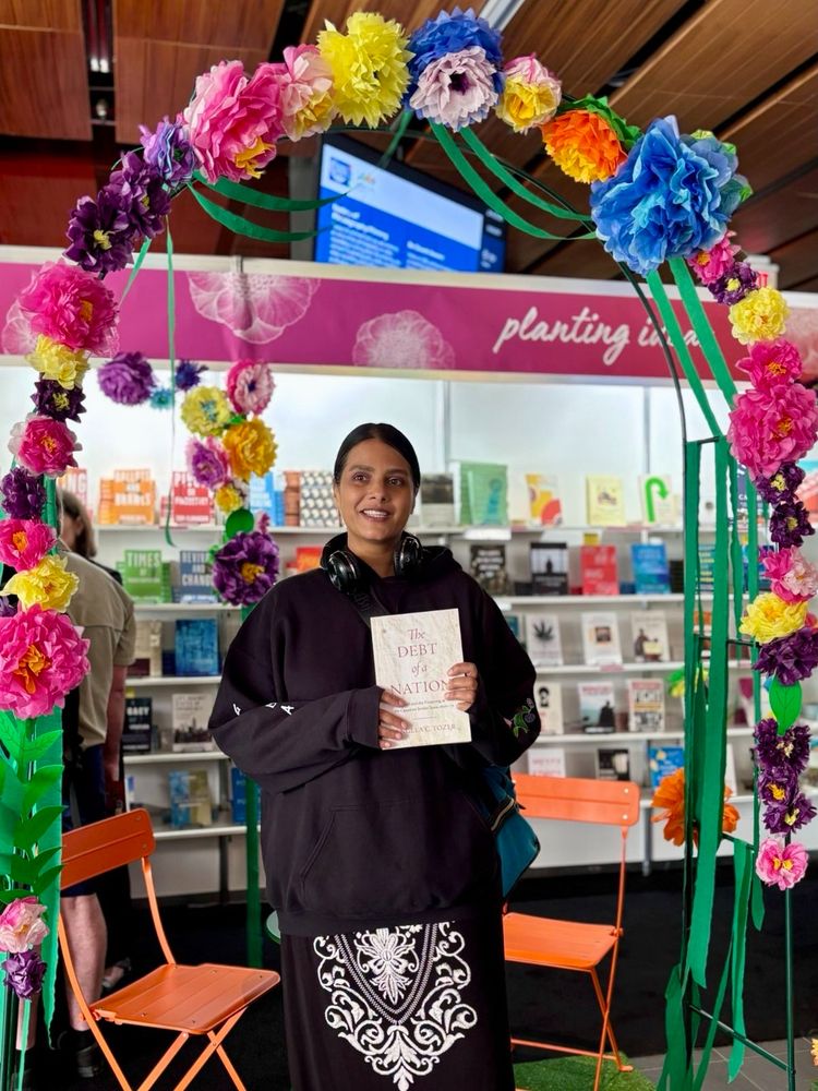 A woman in a black hoodie wearing a black skirt with a white floral pattern, standing under an archway with various artificial flowers holding a book titled "The Debt of a Nation." Behind her is a display with a variety of out of focus books.  
