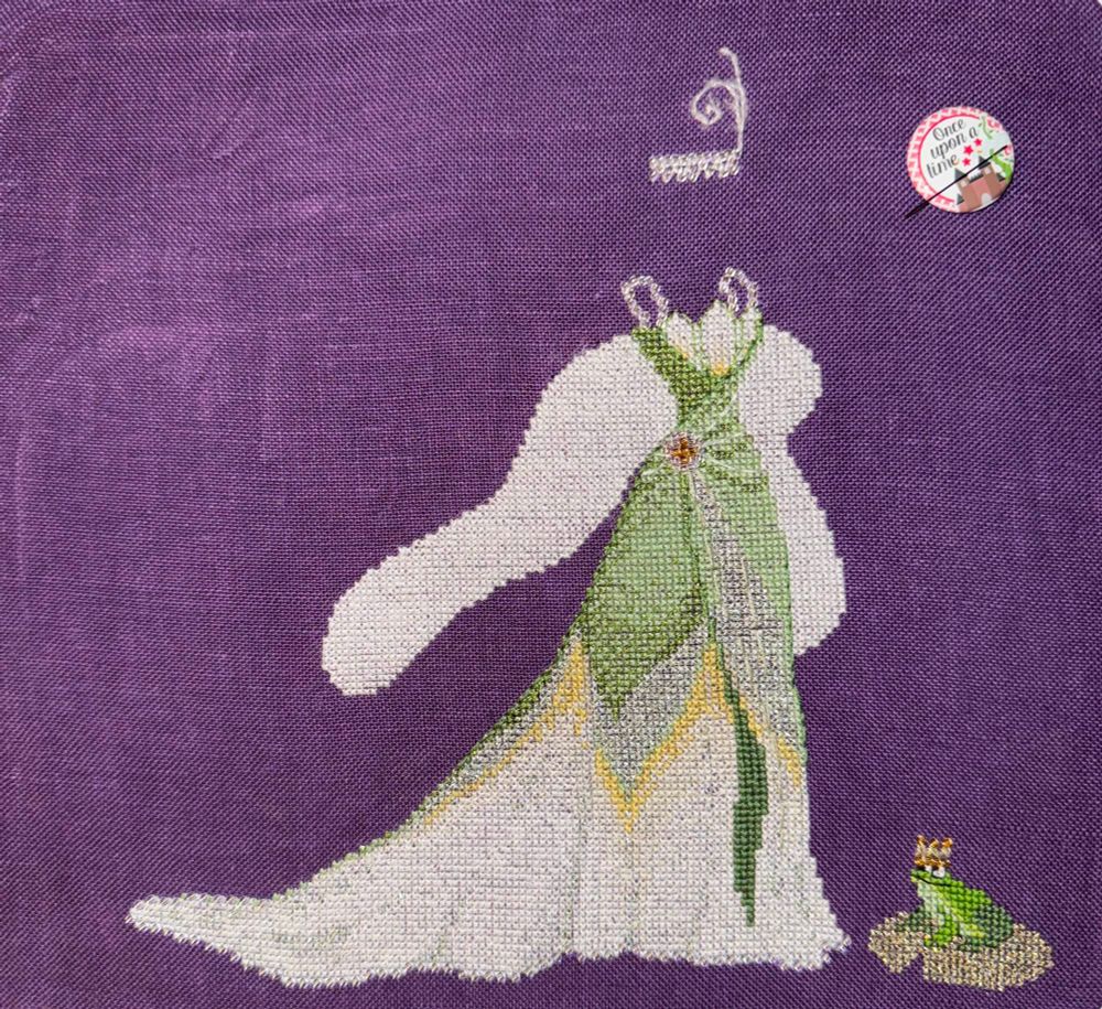 Image: On a dark purple fabric, the green bodice with a white lily breast plate and white feather boa are stitched. The dress makes the shape of an upside down water lily, with green fading into yellow and then into white. A frog sits on a lily pad to the right of the dress. 