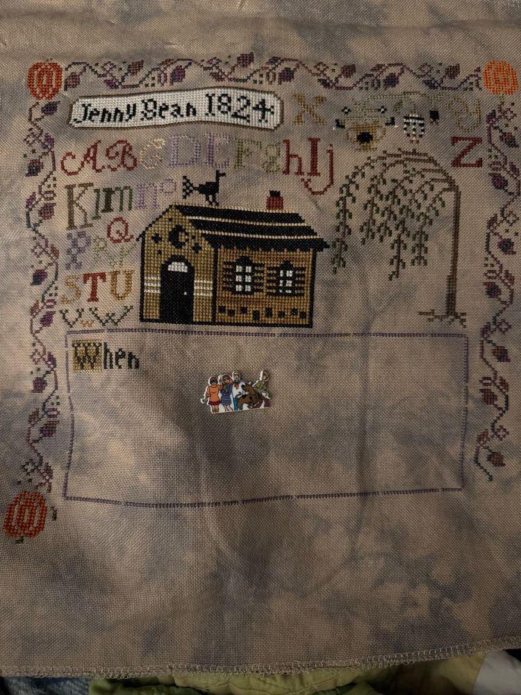 Image Description: On a tan and black mottled fabric, a tan house with black accents has been stitched. The alphabet had been stitched in shades of red, green, brown and purple. Above the alphabet in a box “Jenny Bean 1824” is stitched. A border of vines in shades or purple, grey and brown extend from an orange pumpkin. A potted plant sits between the X and YZ. Below the plant and to the right of the house is a weeping willow tree. Beneath these is a textbox that begins with “When”