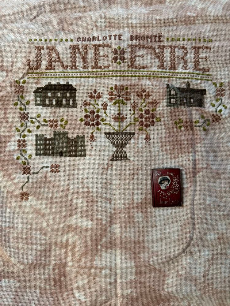  on a mottled pink brown fabric, several green and mauve lines are stitched. The top features Charlotte Brontë’s name and the title “Jane Eyre.” Below that, in the center, a vase with mauve flowers has been stitched. The flowers are wilting.  To their left a manor house is stitched with a floral border. To their right a school has been stitched with a similar border. Brow the manor house is Thonrfield Hall