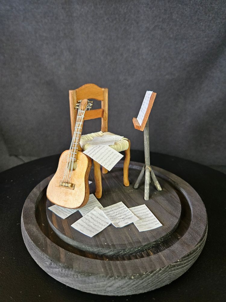 Miniatur in 1:12
Auf einer runden Holzplatte steht ein Stuhl mit Korbgeflecht, an dem Stuhl lehnt eine Akustikgitarre, davor steht ein Notenständer. Auf dem Stuhl und auf dem Boden liegen Notenblätter. Die Miniatur ist komplett handgefertigt 
