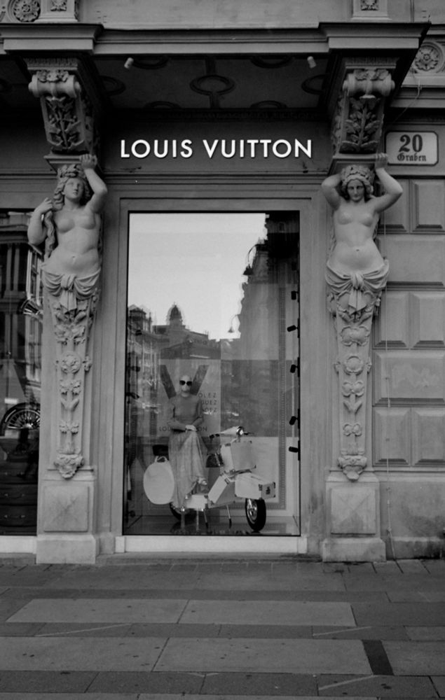 Black-and-white photo. Shopwindow displaying a female mannequin wearing a skirt, a long sleeved t-shirt  and sunglasses. The mannequin is seated on a vespa. In the window some buildings are reflected. The window is framed by two caryatids. Above the window you can read Louis Vuitton in capital letters. Next to the right caryatid the address sign reads the number 20 below which the street name „Graben“ is written.