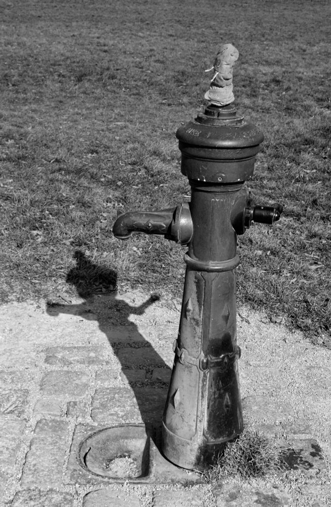 Blackandwhite photo. A water hydrant is casting its shadow onto the cobbled ground. On top of the hydrant a children’s mitten is placed like a hat.