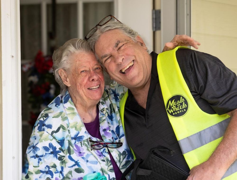Two people smiling at the doorway, one wearing a Meals on Wheels People volunteer vest.