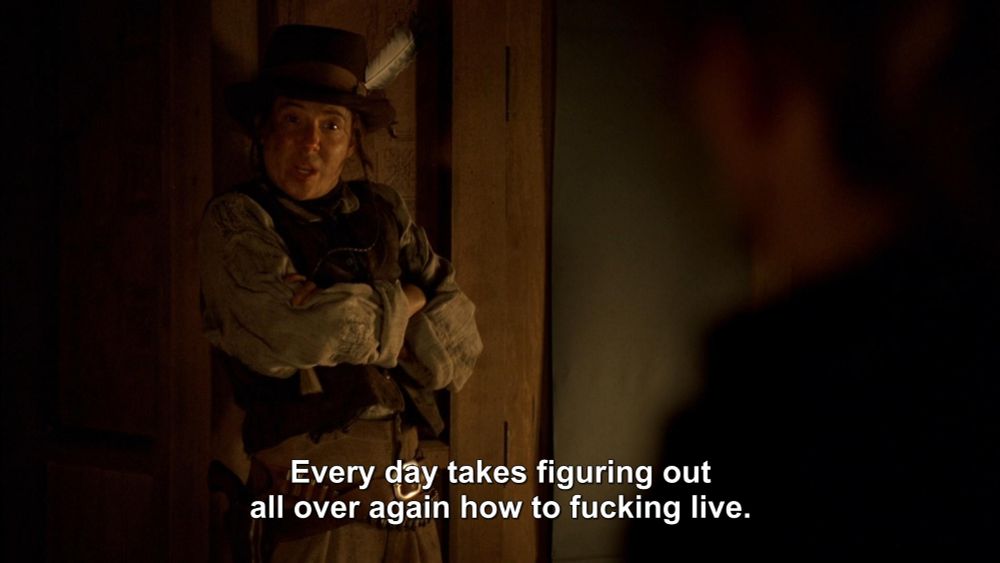 A white woman dressed in trousers, a blouse and a vest with a hat with a feather in it is leaning against a wall or a door with her arms crossed. The text (her dialogue) reads, "Every day takes figuring out all over again how to fucking live."