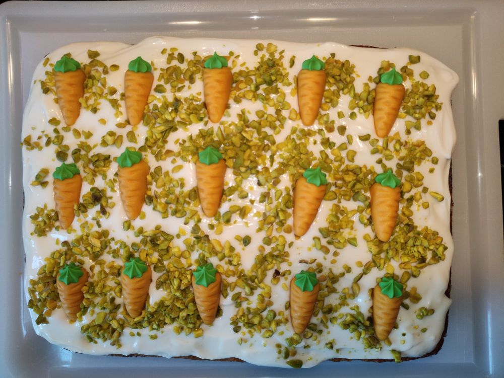 Karottenkuchen auf weißem Kunststoffbrettchen von oben fotografiert.
Weißes Frosting mit gehackten Pistazien und Marzipan Karotten.