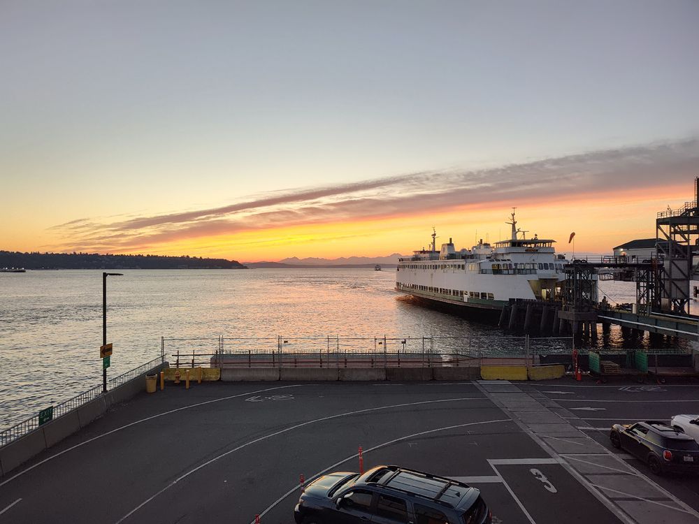 Empty ferry terminal parking lot at sunset with white passenger ferry docked at pier, warm orange sky reflecting on calm water, Olympic mountains in distance