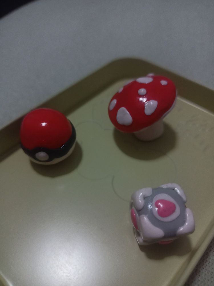 Figuras pequeñas de cerámica polimérica del tamaño de una moneda cada una. Respectivamente una seta, una pokeball y un cubo de compañía de Portal
