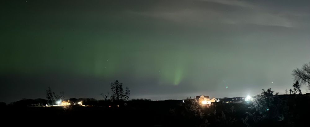 The Northern Lights curling above a couple of rural houses, green ribbons visible through thin wispy clouds. 