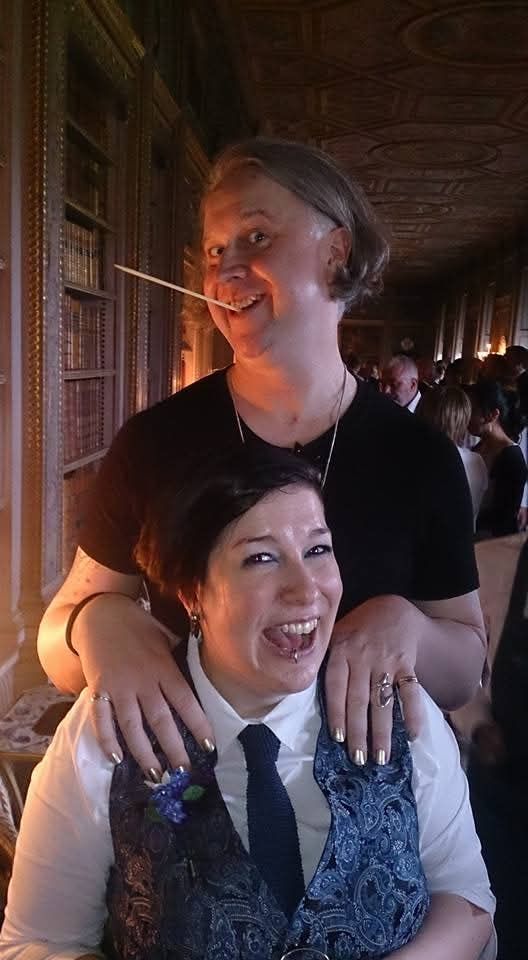 A very tall femme non-binary person in a black short-sleeved jumpsuit and with grey hair cut in a bob, is pulling a silly grin at the camera with a wooden skewer gripped between their teeth. They are stood directly behind me with their hands on my shoulders. I am a short non-binary person in a white shirt, navy tie and paisley blue waistcoat. I have black hair in an asymmetric short style,and I'm giving a wide mouthed smile at the camera. We're both a bit shiny and loose thanks to the copious prosecco being served at our friends' wedding.