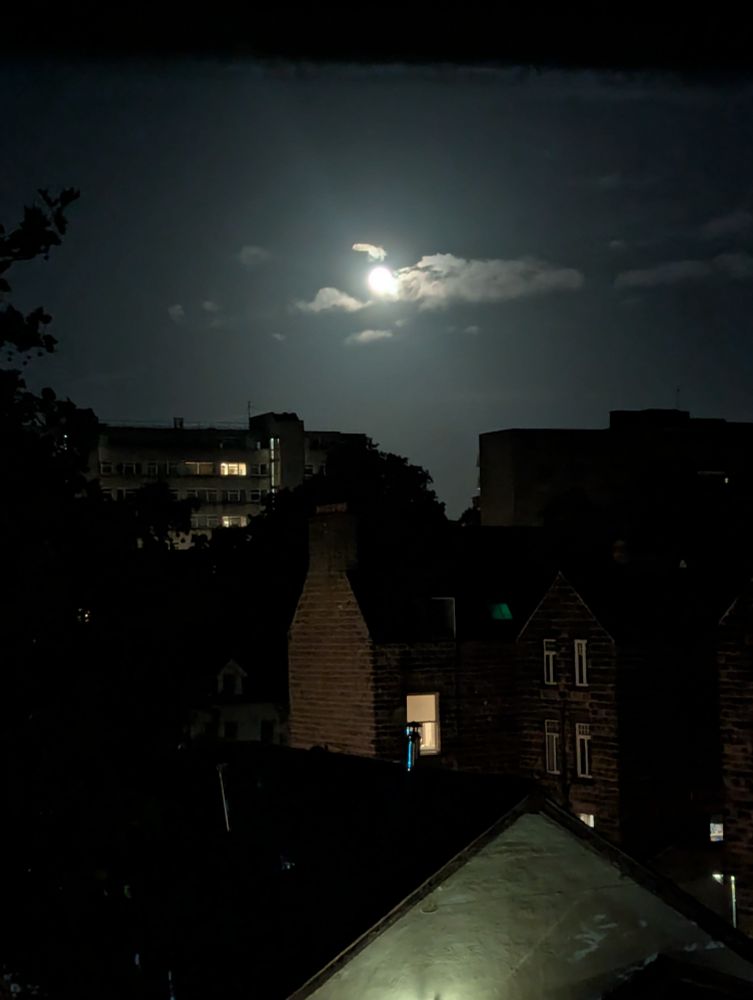 Photo over the roofs of some buildings, with the moon very bright and low in the sky and some clouds