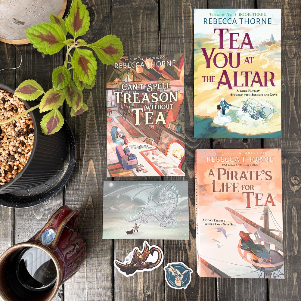 An image of 3 books (Can't Spell Treason Without Tea, A Pirate's Life for Tea, and Tea You at the Altar), framed by a postcard of a dragon image and 2 vinyl stickers (of a griffon and a dragon). Surrounding the items is a mug and some plants. 