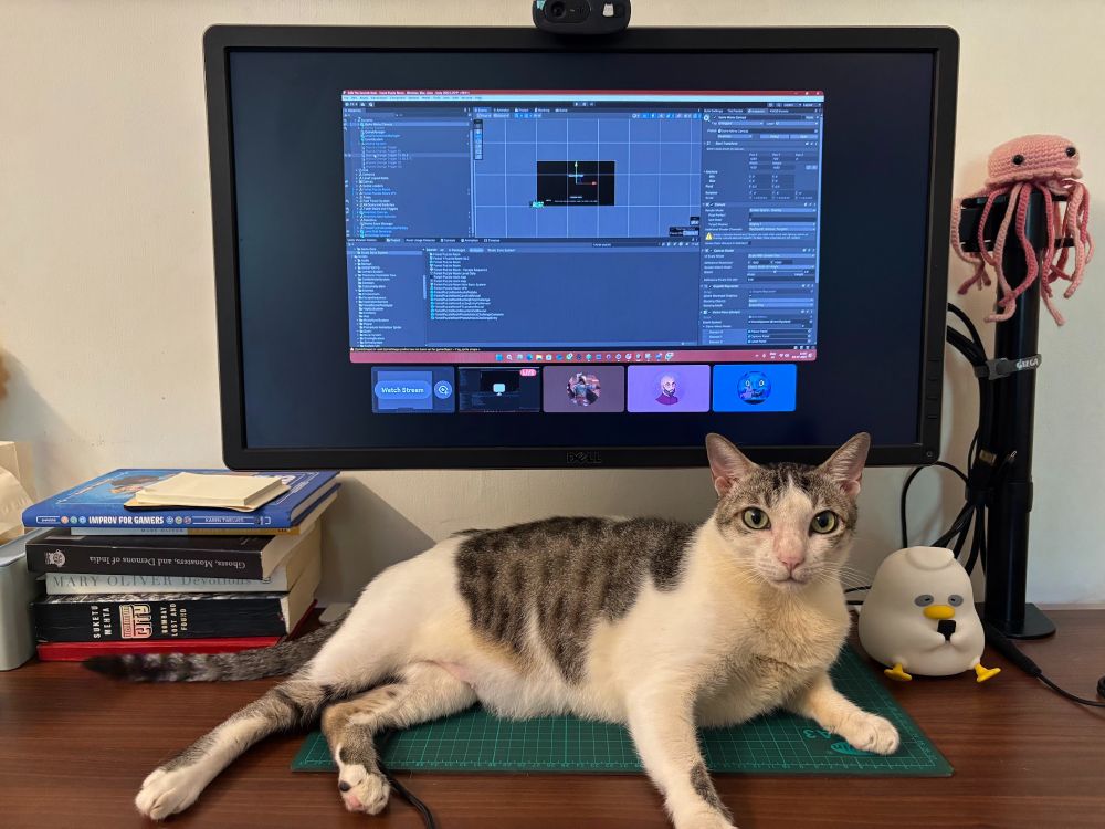 cat looking at camera while under a pc monitor. the monitor has Unity open.