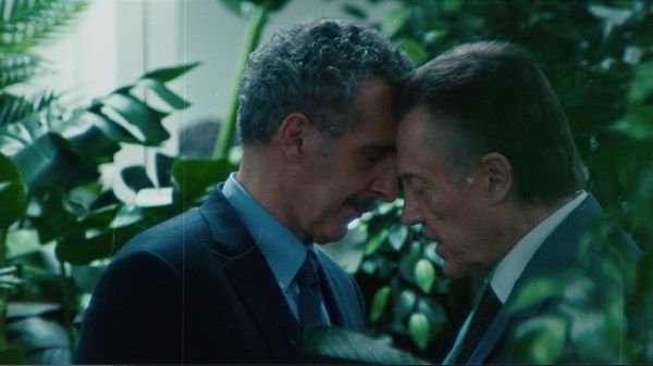 Famous older white male actors John Turturro and Christopher Walken lean into each other, touching foreheads, while standing amid a bunch of green plants 
