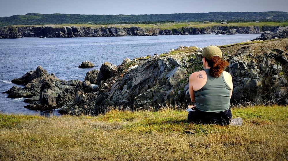 A woman with red hair a green vest looks out over the water from a cliffside.