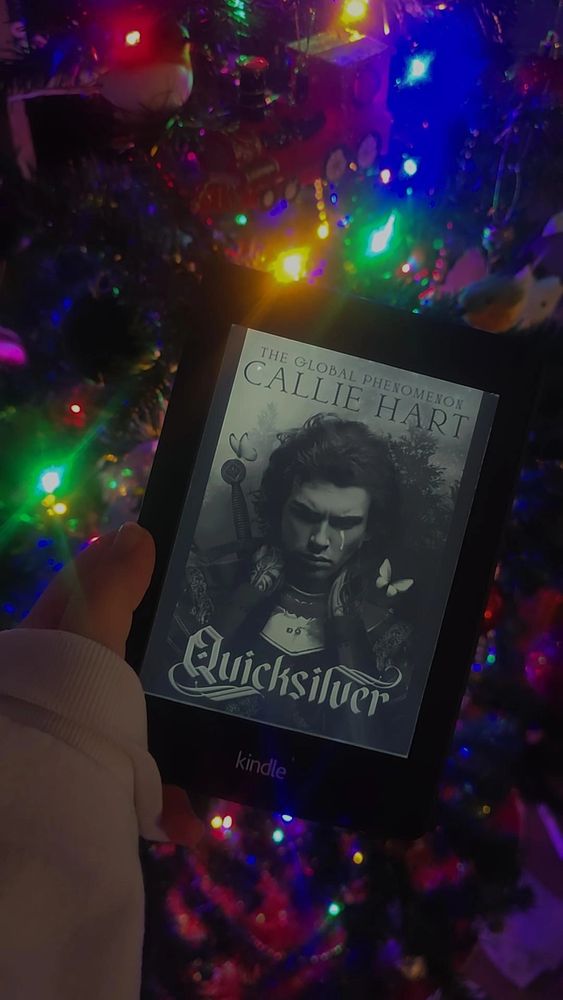 A Kindle with the black and white image of a man with butterflies and a sword as the front cover of Quicksilver by Callie Hart. A Christmas tree lit with colourful lights is in the background.