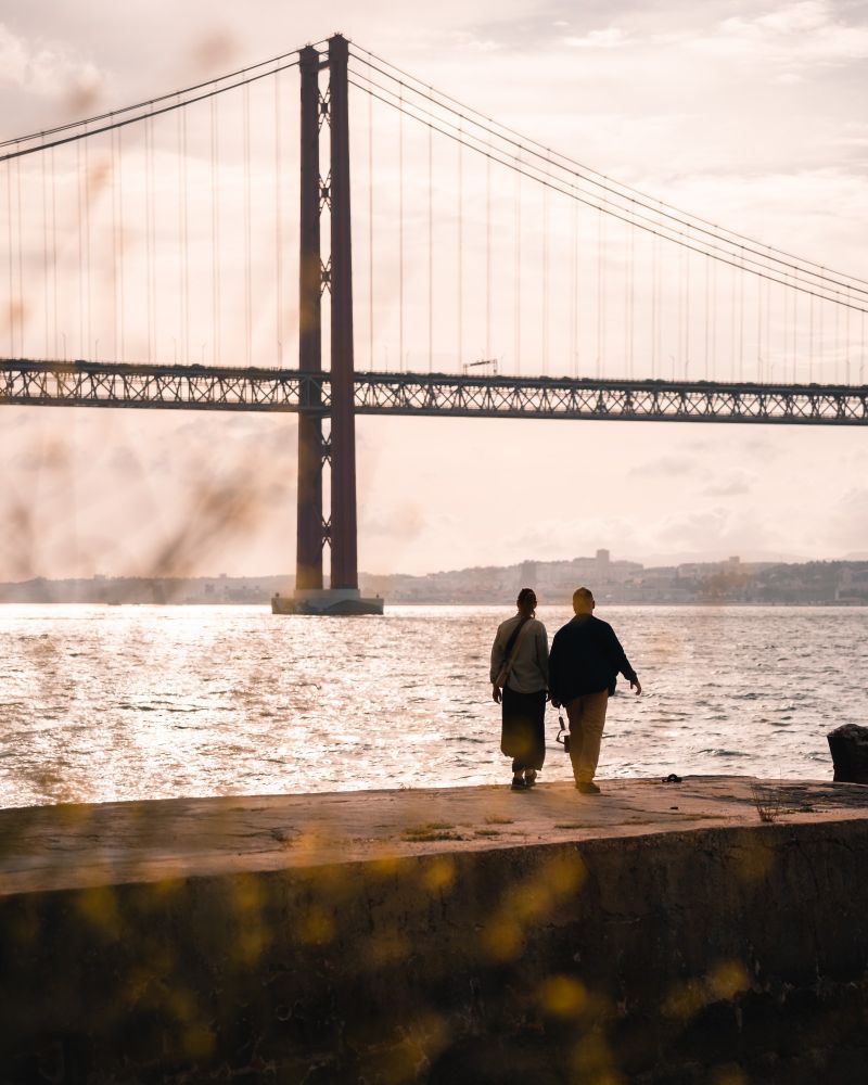 Sunset photo of a couple walking by the river with ponte 25 de abril in Lisbon in the background