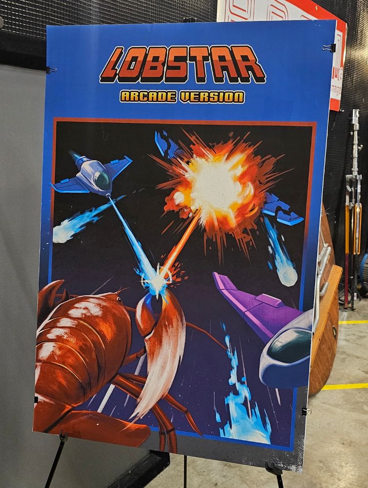 A poster titled "Lobstar Arcade Version", depicting a lobster in space as it punches a projectile into a spaceship. Two smaller spaceships can be seen on either side of the creature.
