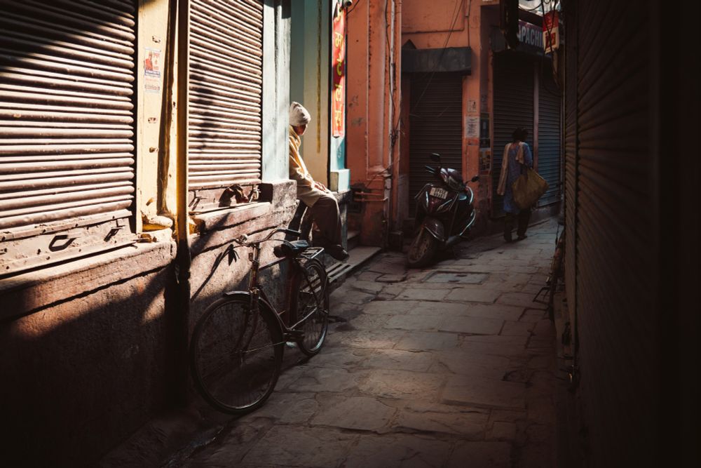 A narrow street in Varanasi, India. Shop shutters line each side of the image and lead you towards a lone figure sitting upon a step in the centre of the frame. The images is mostly dark apart from a bright area of sunlight that has penetrated the gloomy narrow lane.