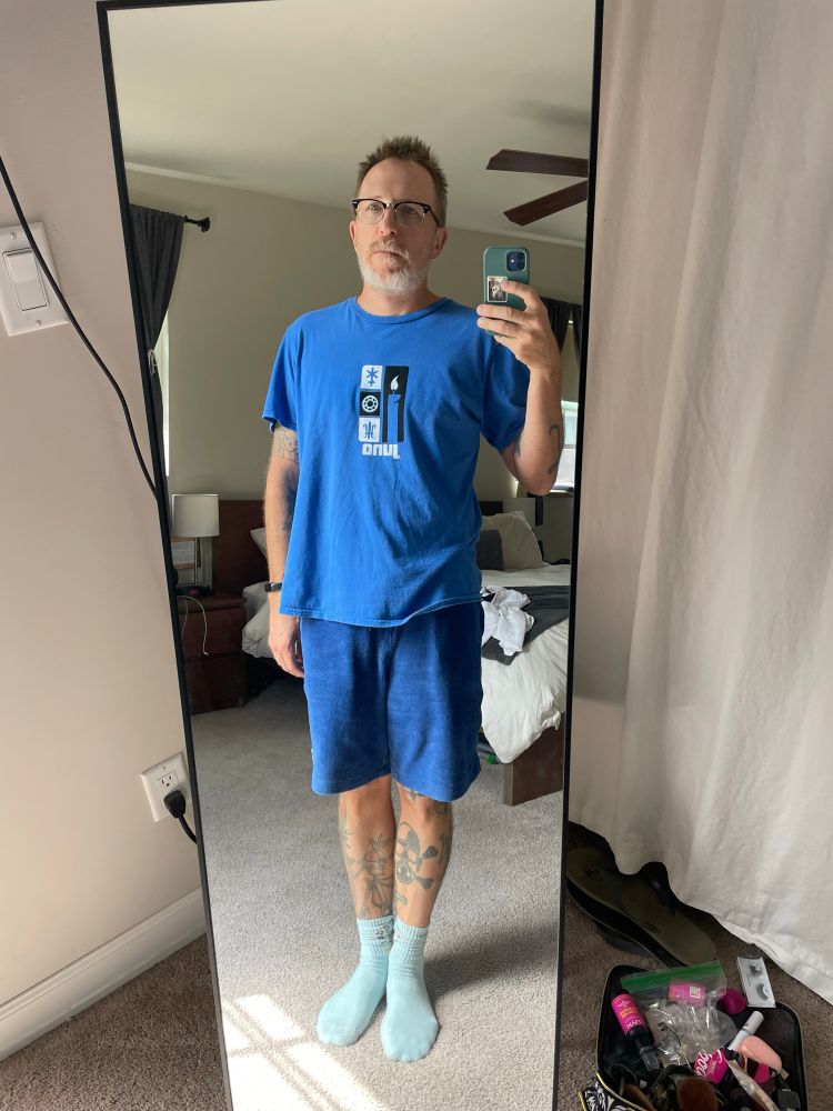 A man with tattoos, glasses and a grey beard wearing a blue shirt, blue shorts, and blue socks, takes a mirror selfie. 