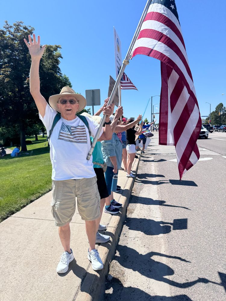 An older man in a white tshirt with an American flag on it, khaki shorts, and a hat and sunglasses waves at the camera while holding an American flag at the No Kings rally in Denver. 