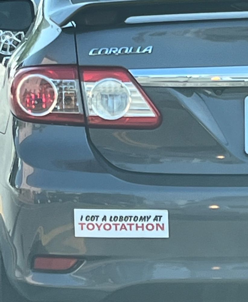 The back left bumper of a grey Toyota Corolla with a white bumper sticker that reads, “I GOT A LOBOTOMY AT TOYOTATHON”