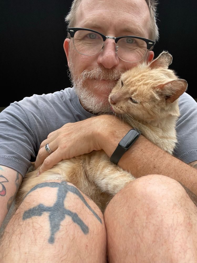 Older man in glasses, with tattoos and a grey beard cuddles an orange cat.