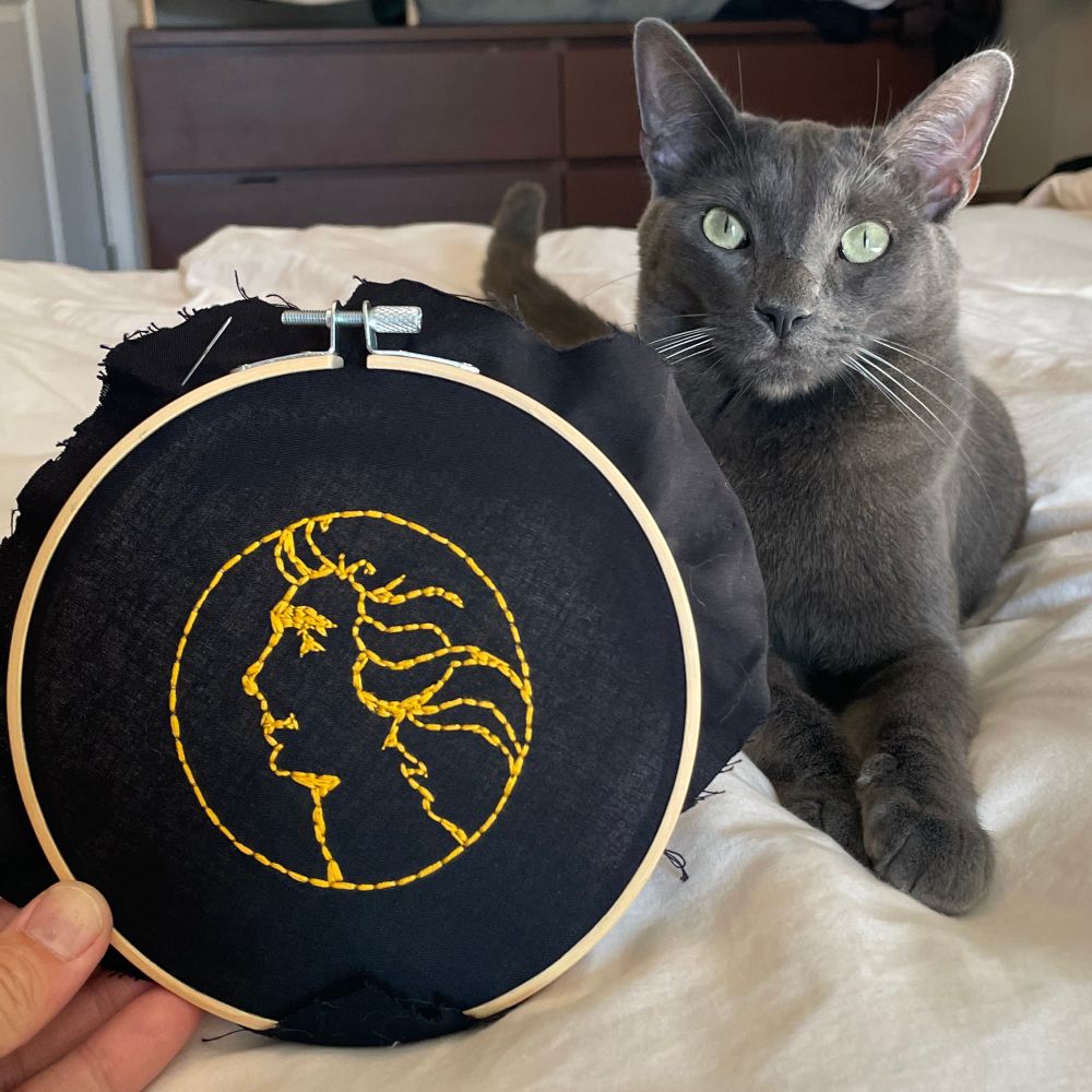 A grey cat sits next to an embroidery of Barry Manilow, made with gold floss on black fabric. 